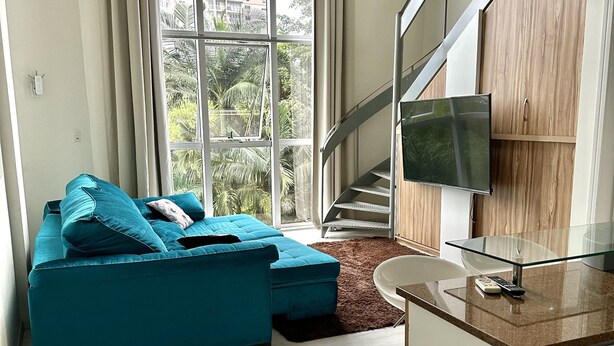 A modern living room with a teal sofa, a glass coffee table, and a TV mounted on a wooden cabinet. A staircase is visible in the background. - Apartamento 206 Saint Sebastian Flat Duo (Jaragua do Sul)