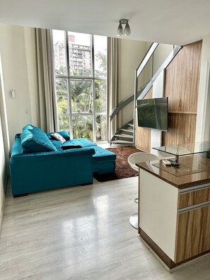 Een moderne woonkamer met een blauwgroene bank, een glazen salontafel en een televisie bevestigd aan een houten kast. Een trap is op de achtergrond zichtbaar.