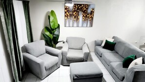 Ein modernes Wohnzimmer mit einer grauen Sofagarnitur, einem grauen Sessel und einem Couchtisch. Eine große, grüne Pflanze und ein Kunstwerk befinden sich über der Couch.