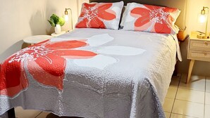 Ein ordentlich bezogenes Bett mit einem gemusterten Überwurf, ein Nachttisch mit Lampe und ein kleiner Blumentopf.