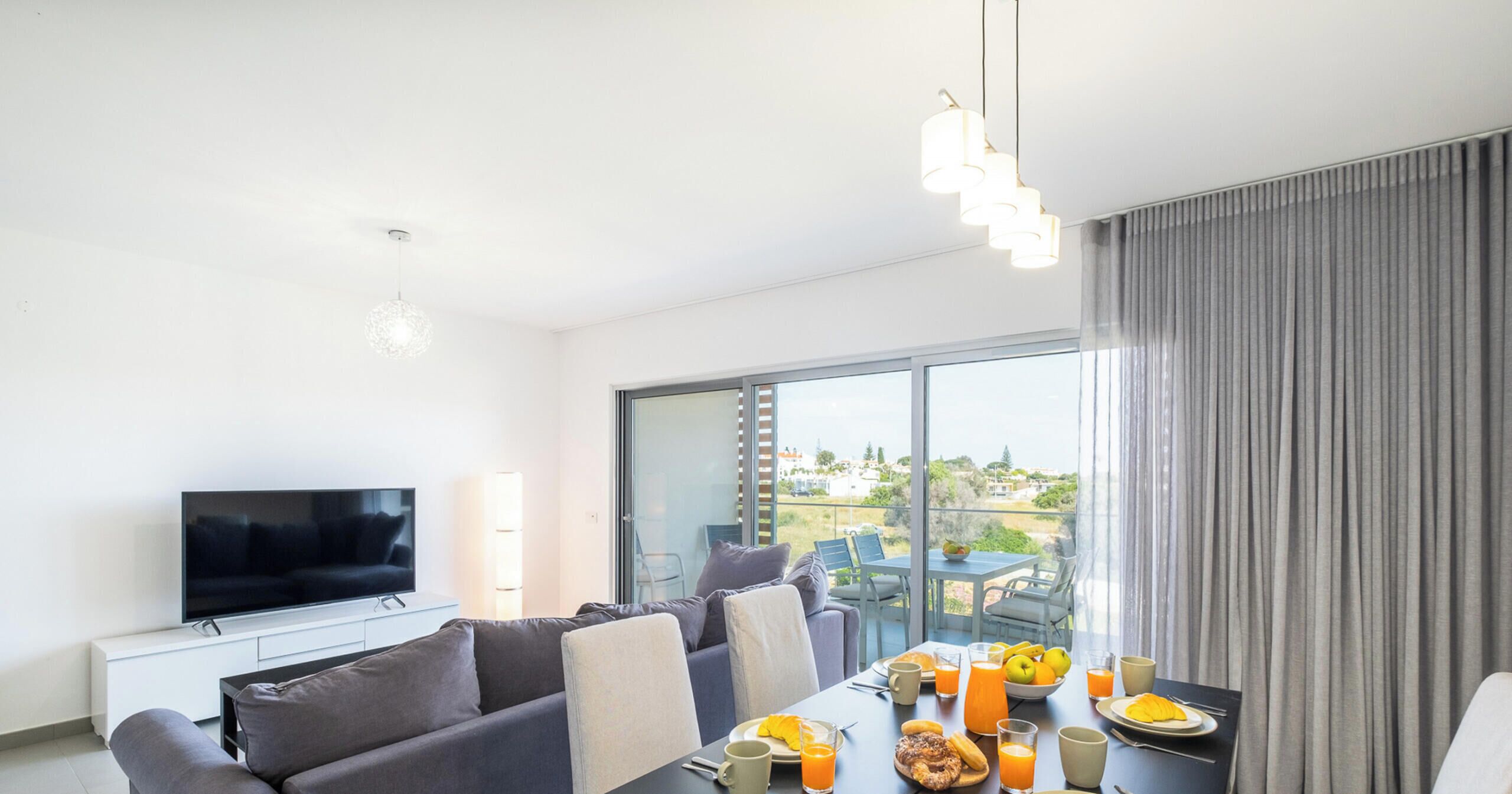 A modern living room with a flat-screen TV, grey sofa, dining table with breakfast items, and a large window with a view of greenery.