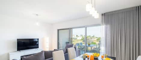 A modern living room with a flat-screen TV, grey sofa, dining table with breakfast items, and a large window with a view of greenery.