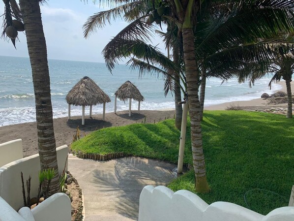 Una playa con sombrillas de paja, palmeras y un camino de hormigón que conduce a la orilla.
