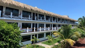 Een hotel met twee verdiepingen, een rieten dak, meerdere balkons en een tuin met palmbomen.