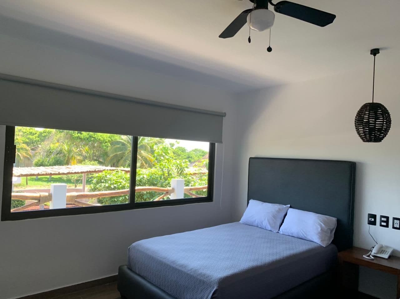A bedroom with a bed, a nightstand, a phone, a ceiling fan, and a window with a view of greenery.