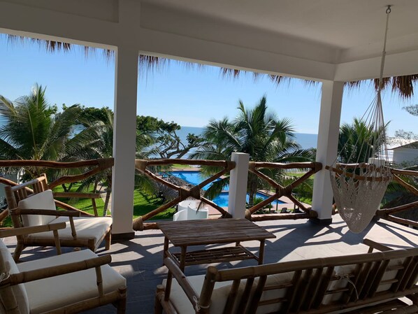 A covered outdoor seating area with bamboo furniture, a hammock, and a view of a pool and palm trees.