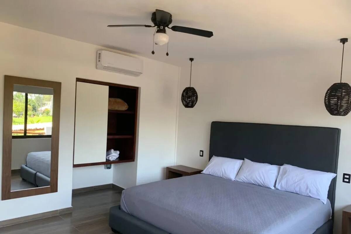 Un dormitorio con cama, ventilador de techo, espejo, armario y ventana.