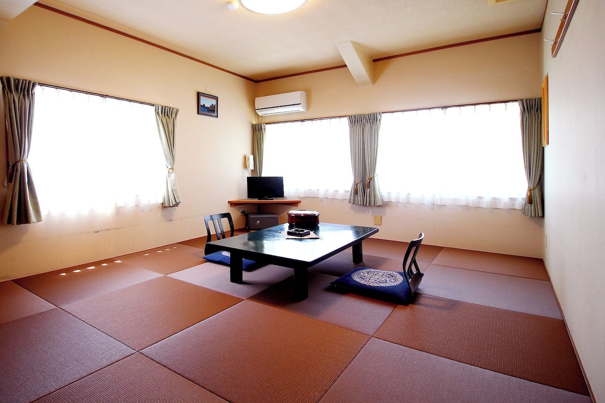 A room with tatami mats, a table with chairs, a television, and large windows with curtains.