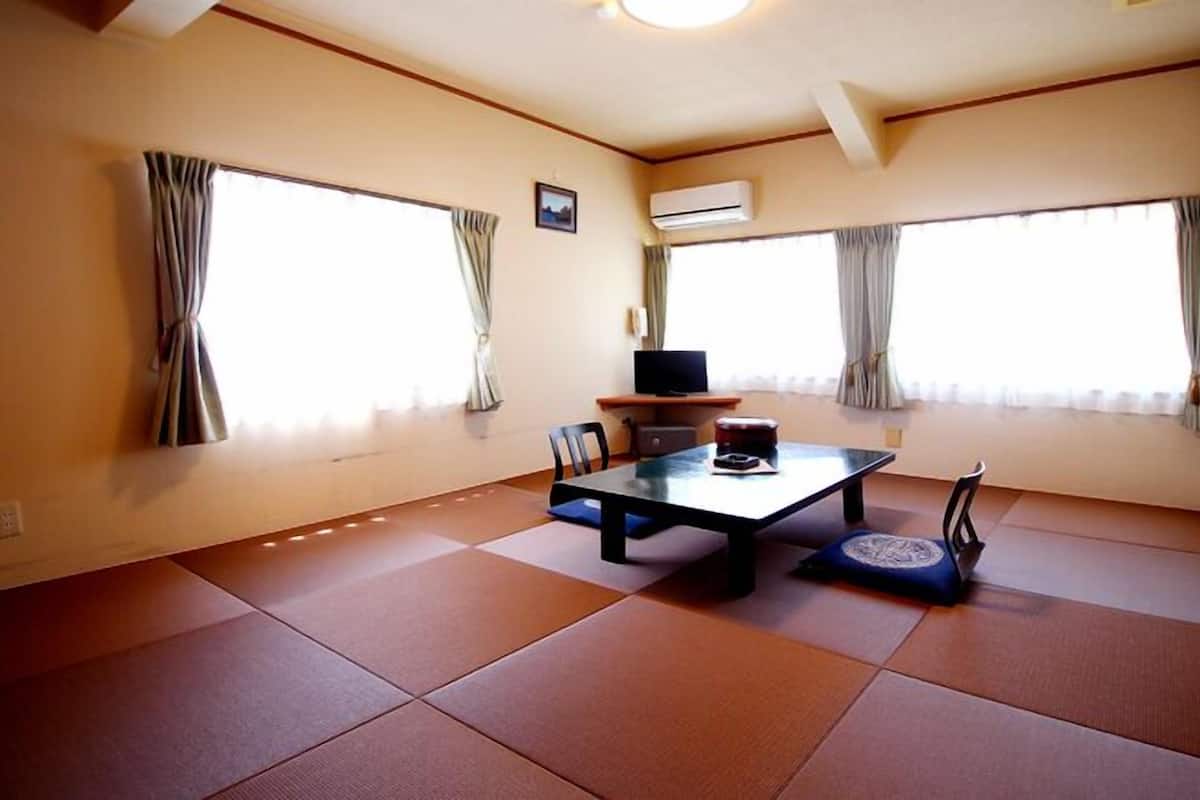 A room with tatami flooring, a low table with chairs, a television on a stand, and large windows with curtains.