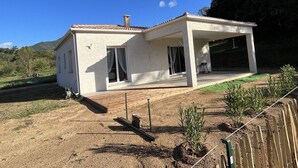 Una casa de un piso con un porche techado, una chimenea visible y un patio cercado con plantas jóvenes.