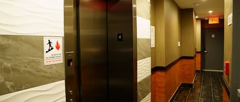 Een lift met een brandveiligheidssymbool, een gang met een marmeren wand en een vluchtwegbord.