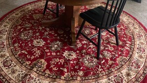 Een eethoek met een ronde houten tafel, twee zwarte stoelen, een tapijt met een rood en beige patroon, en een potplant met rode bloemen.