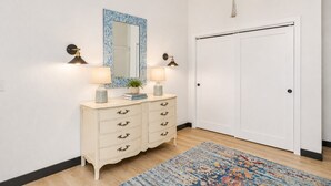 A room with a white door, a cream-colored dresser with multiple drawers, two wall-mounted lamps, a mirror with a blue frame, and a patterned rug on the floor.