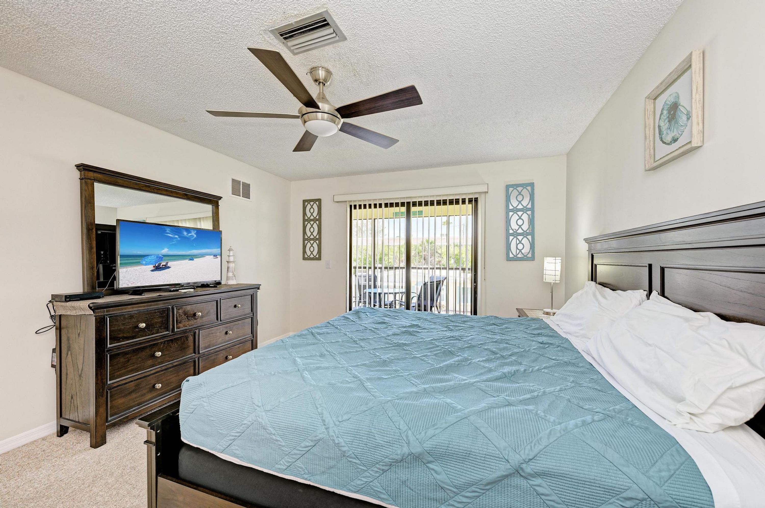 Quarto com cama, penteadeira, ventilador de teto, janela com grades e um quadro na parede.