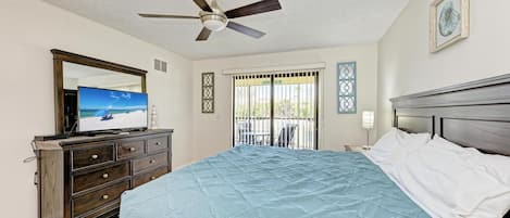 Quarto com cama, penteadeira, ventilador de teto, janela com grades e um quadro na parede.