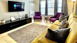 Olohuoneessa on taulutelevisio, kaksi violettia nojatuolia, keltainen sohva ja pieni pyöreä pöytä.