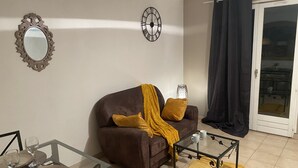 Una sala de estar con un sofá marrón, una mesa de centro de cristal, una mesa de comedor con platos y vasos, un espejo y un reloj en la pared.