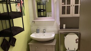 Un baño pequeño con un lavamanos blanco, un inodoro y un espejo sobre el lavamanos. Hay un perchero con toallas y una pequeña estantería con artículos de aseo.