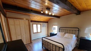A bedroom with a wooden ceiling, a metal bed frame, a bedside table with a lamp, and a wardrobe. -  Carriage House in Old Saratoga (Schuylerville)