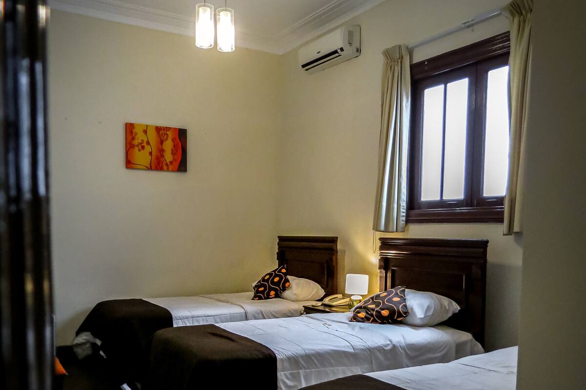 Ein Hotelzimmer mit zwei Betten, einem Nachttisch, einer Lampe, einer Klimaanlage, einem Fenster mit Vorhängen und einem Bild an der Wand.