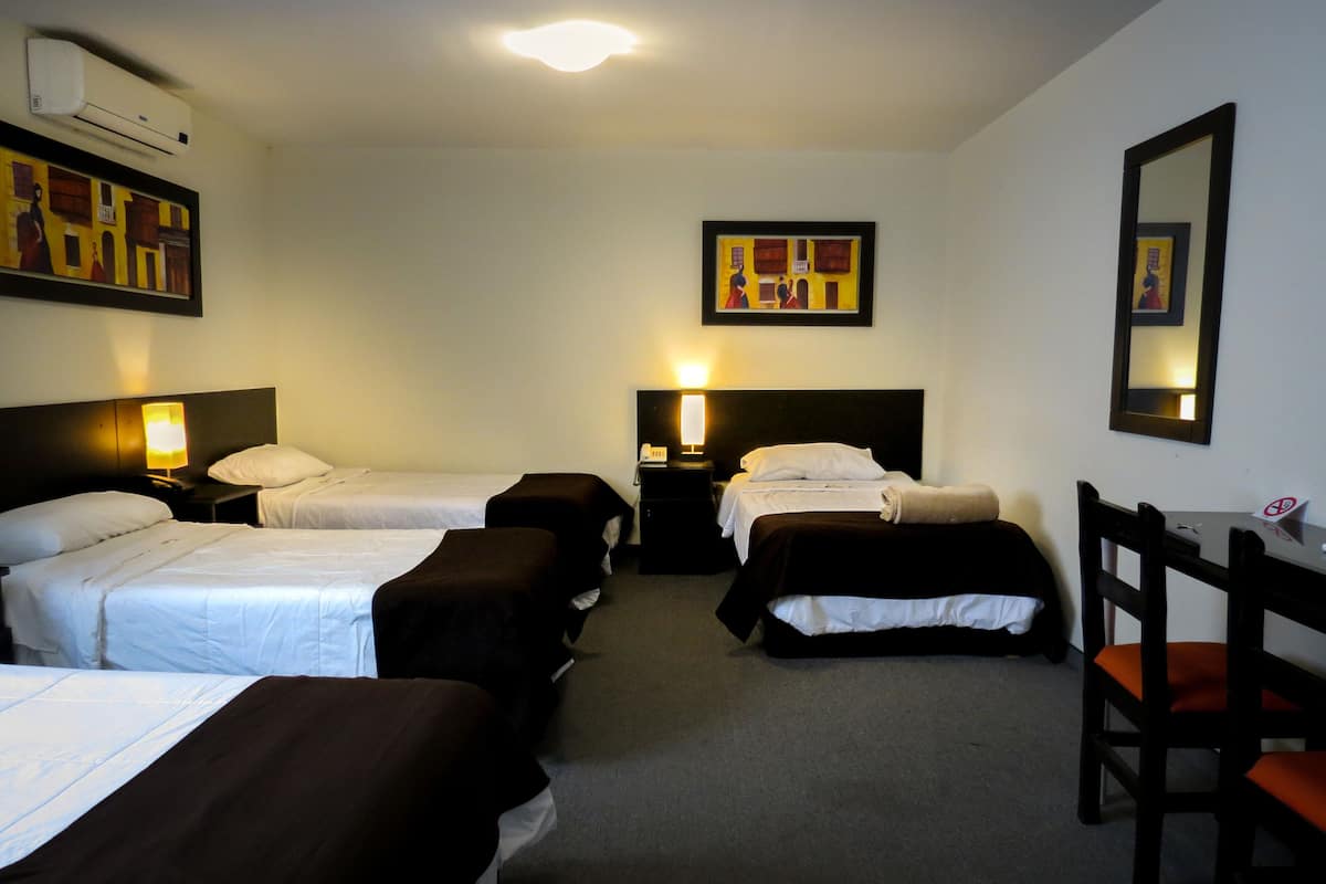 Ein Hotelzimmer mit drei Einzelbetten, einem Schreibtisch und einem Stuhl. Zwei Wandlampen und eine Deckenleuchte sind vorhanden.