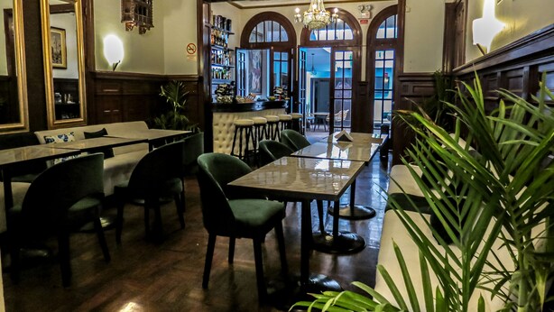 Un restaurante con zona de bar, varias mesas y sillas. Hay plantas en macetas y un candelabro.