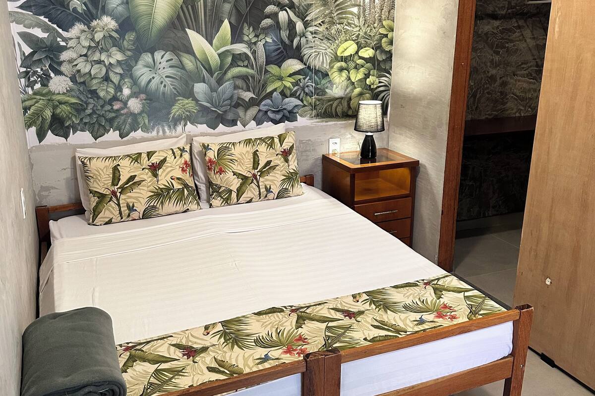 A bedroom with a bed, a nightstand, a lamp, and a tropical-themed wall mural.