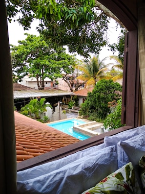 A view from a window of a room with a bed, a pool, and a courtyard with trees and buildings. - Jardim Secreto Guest House - Hostel (Búzios)