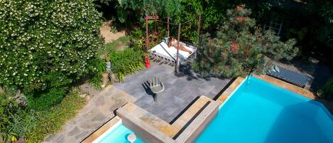 Una piscina con borde de baldosas, rodeada de exuberante vegetación y un pequeño espacio de estar con mesa y sillas.