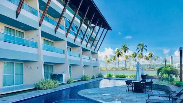 A swimming pool with a curved design, surrounded by a patio area with outdoor furniture. - Villas Do Atlântico (Ipojuca)