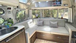 A compact RV interior with a kitchenette, a seating area with cushions, and a window providing a view of greenery.