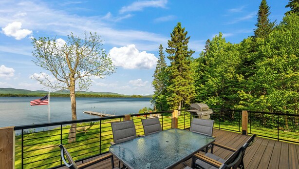 Une terrasse avec une table et des chaises, donnant sur un lac où flotte un drapeau, le tout entouré d’arbres.