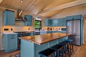 A modern kitchen with wooden ceiling beams, a central island with stools, and stainless steel appliances. - Camp 16 | NEWLY Renovated! Sandy Beach, Private Dock, Mooring, Fire Pit (Rangeley)