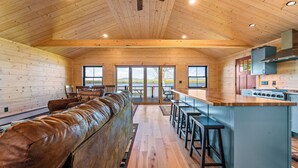 L’intérieur d’une cabane en rondins présente un long canapé en cuir, une cuisine avec un îlot central et de grandes fenêtres.