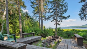 Eine Holzterrasse mit Bänken und einem Feuerkorb, umgeben von Bäumen und Blick auf einen See.