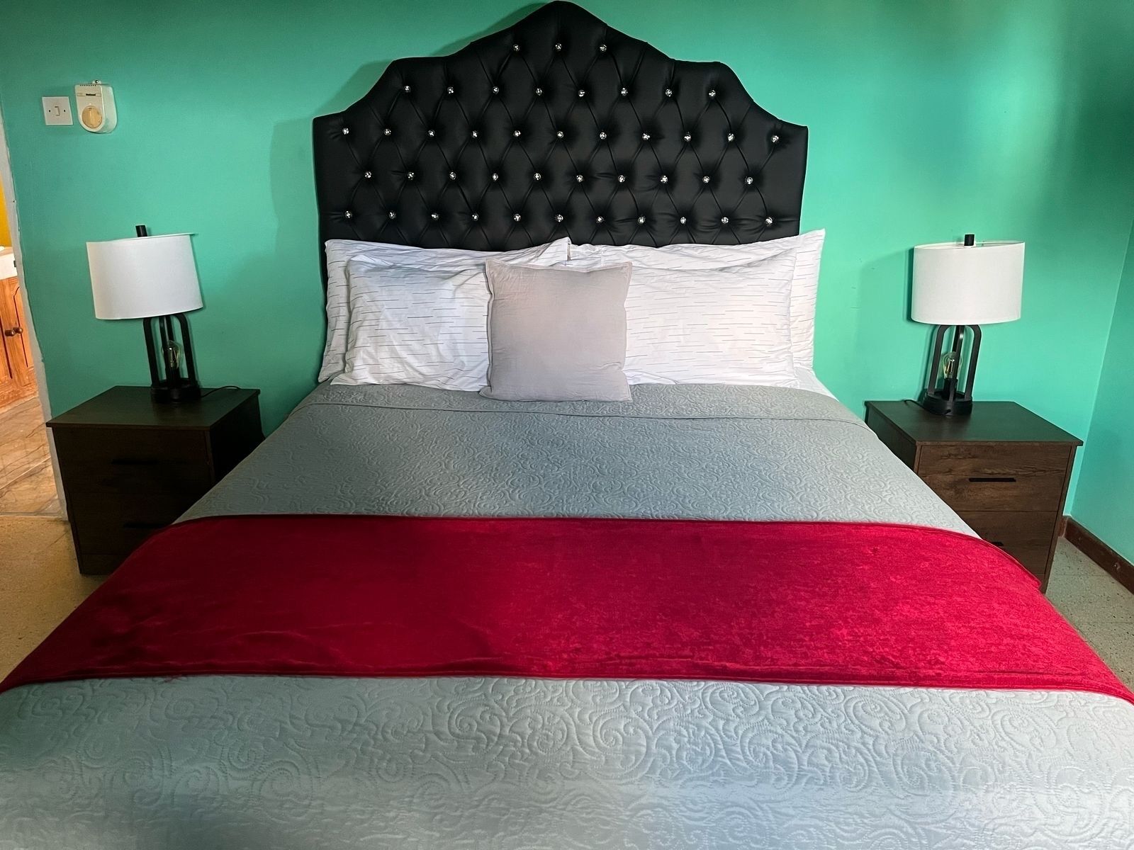 Een bed met een gestoffeerd hoofdeinde, beddengoed in wit en grijs, en een rode plaid. Er staan twee nachtkastjes met lampen.
