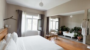 Habitación de hotel con una cama grande, una zona de estar con sillas, una mesita, una bañera y varias plantas en macetas.