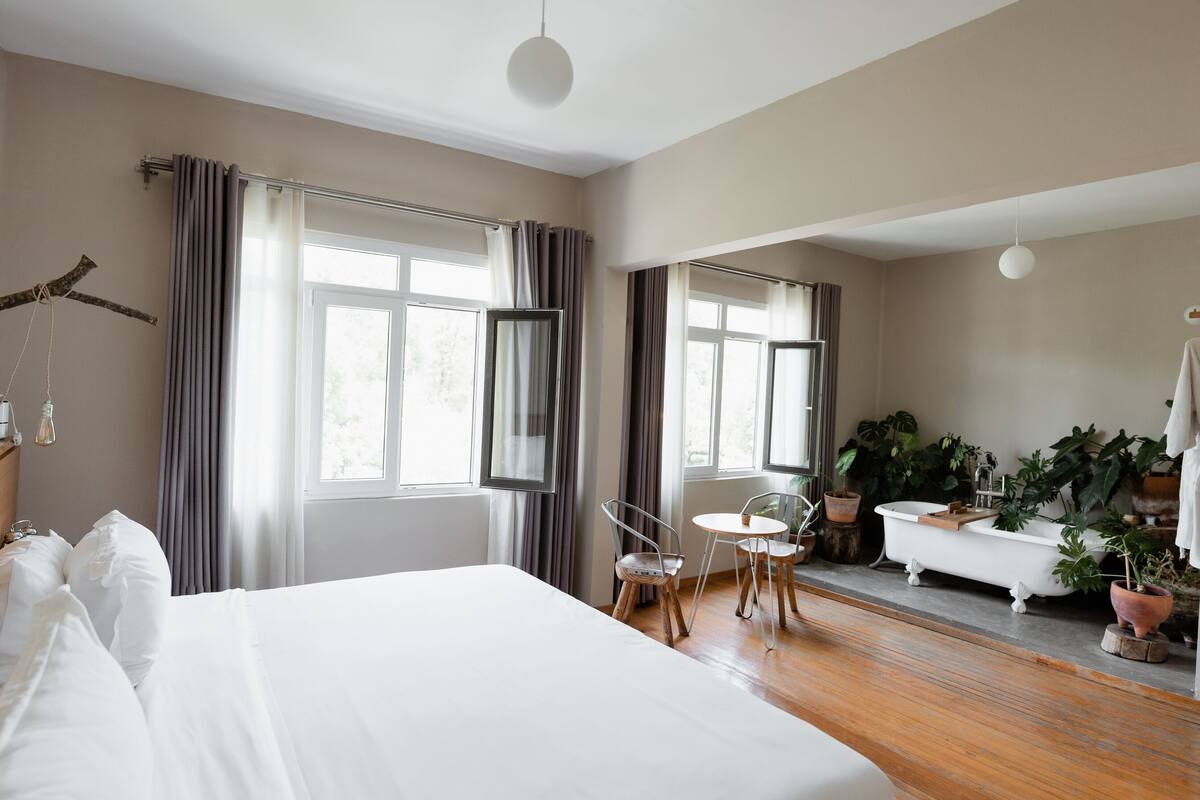 A hotel room with a large bed, a sitting area with chairs, a small table, a bathtub, and several potted plants.