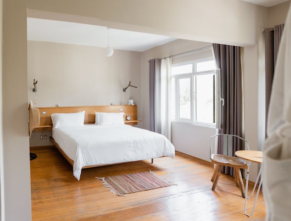 A hotel room with a wooden bed, a small table, a chair, a window with curtains, and a wall-mounted light fixture.