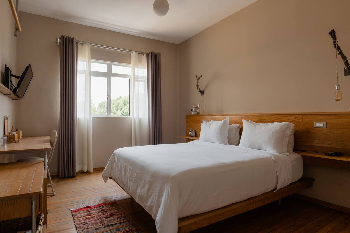 A hotel room with a wooden bed, a desk, a chair, a window with curtains, a wall-mounted light fixture, and a small shelf with a clock and a decorative item.
