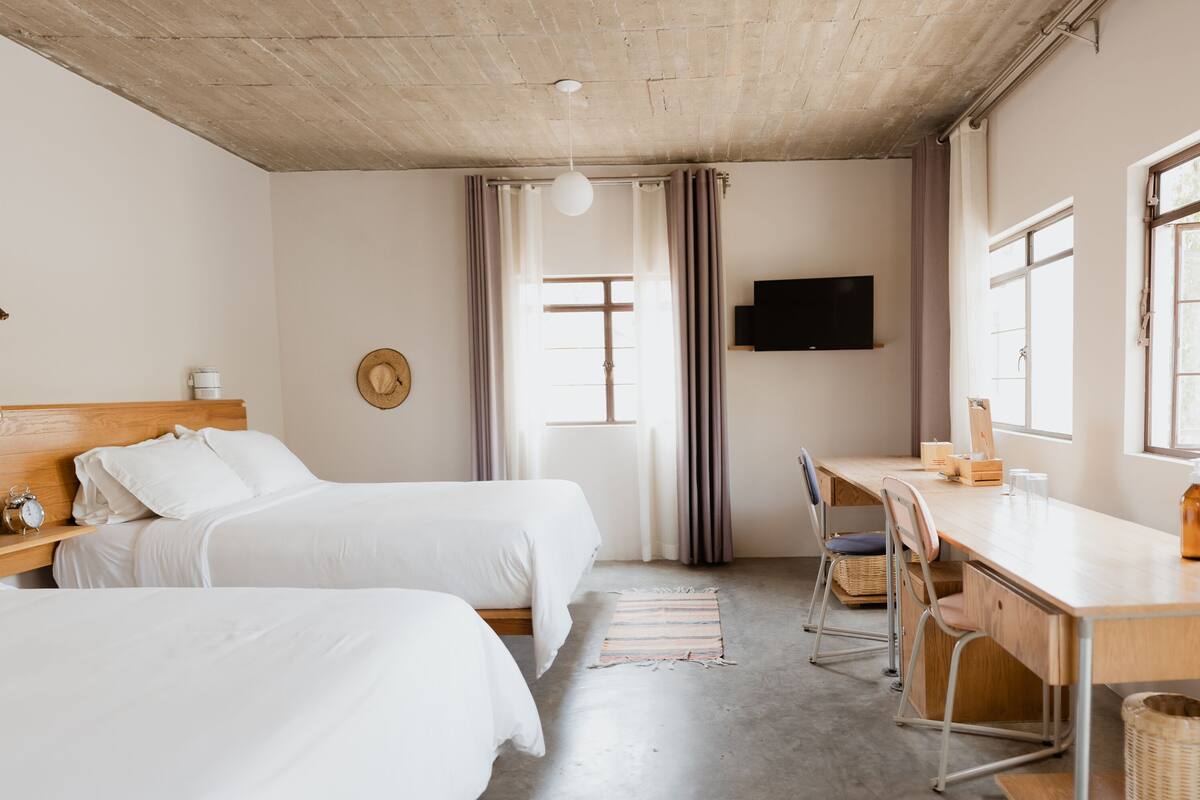 A hotel room with two beds, a desk, a TV, and a large window.