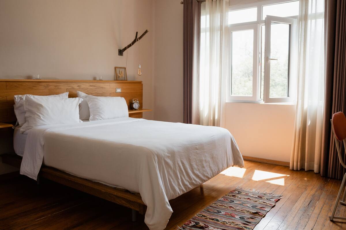 A neatly made bed with white linens, a wooden headboard, a bedside table with a lamp, and a view of the outdoors through a window with curtains.