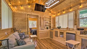A cozy cabin interior with a wooden floor, a sofa with cushions, a small table, a kitchen area with a sink and cabinets, and a large window with blinds. - Grill, Fire Pit: Pet-Friendly Lakeside Cottage! (Lakeside)