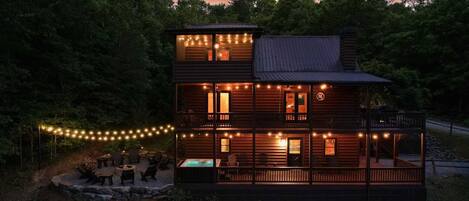 A two-story log cabin with a hot tub and outdoor seating area, surrounded by trees.