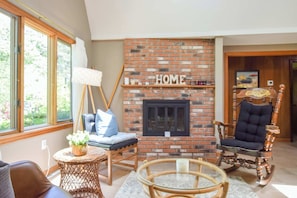 A living room with a brick fireplace, a wooden rocking chair, a glass coffee table, and a wicker side table.