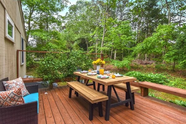 An outdoor deck with a wooden table set for dining, surrounded by benches, and a sofa with patterned cushions. - Central AC 3 Min Walk to Pond! (Marstons Mills)