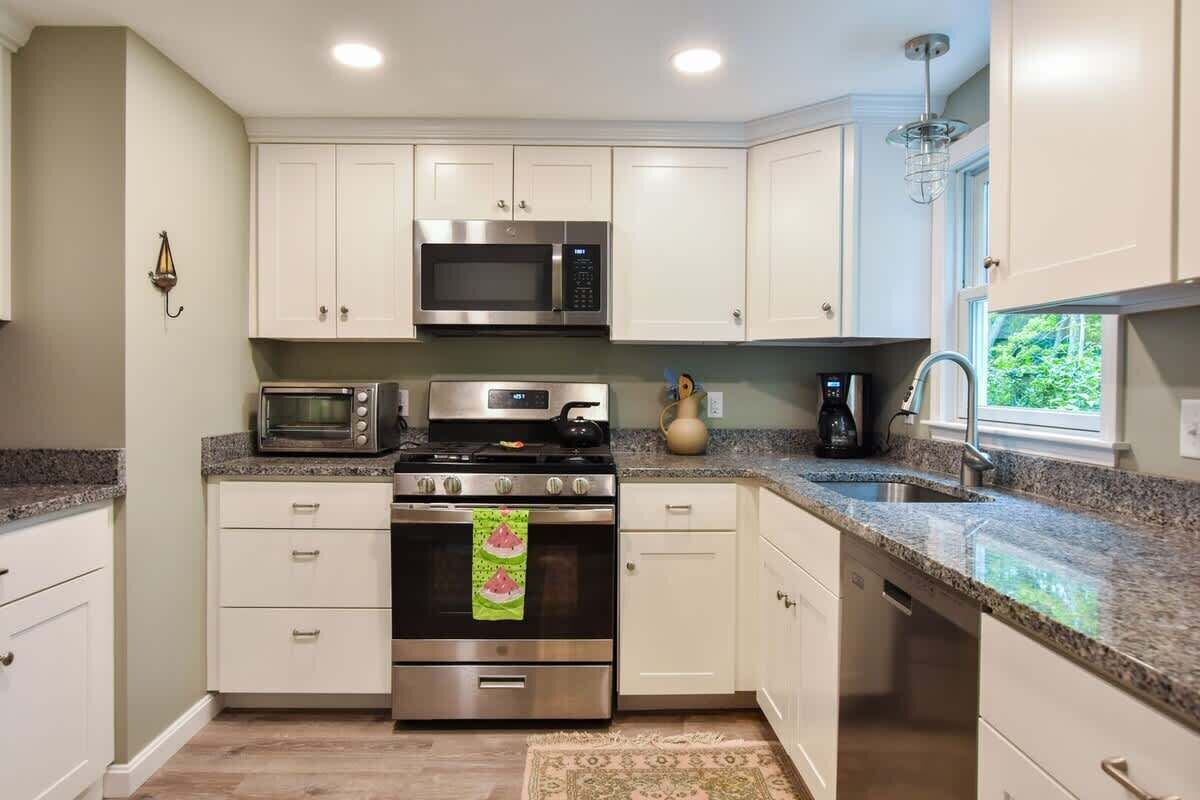 A kitchen with white cabinets, a microwave, an oven, a sink, and a window with a view of greenery.