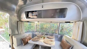 A modern RV interior with a dining area, cushioned seating, and a large window offering a view of greenery. - Chic Airstream Glamper Near Tybee & Savannah (Savannah)