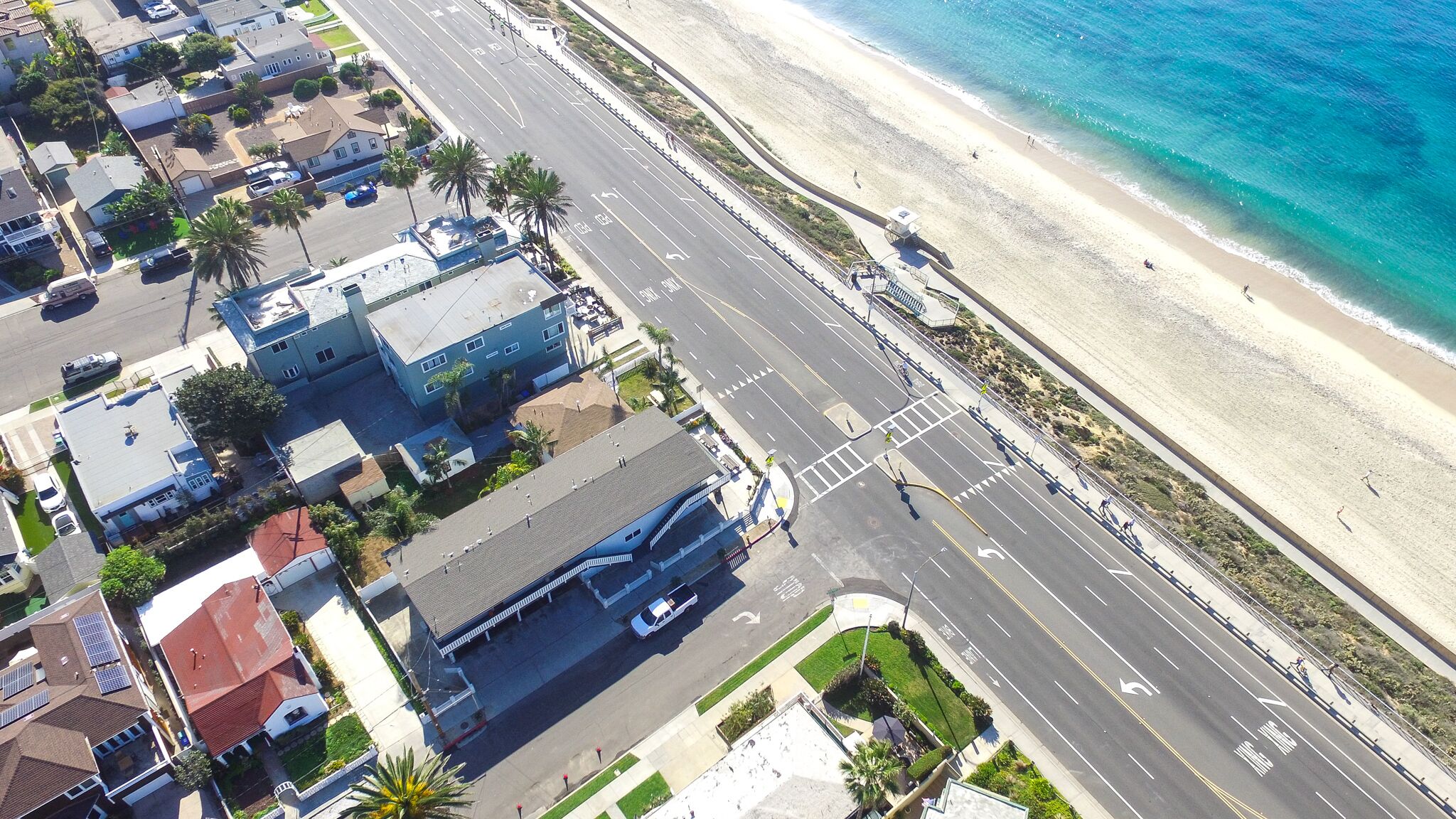 An aerial view of a coastal street with buildings, a beach, and a road with vehicles.