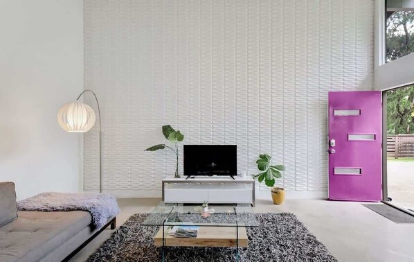 A modern living room with a flat-screen TV on a white console, a grey sofa, a glass coffee table, and a large plant. - Plum Perfect | Cozy SoCo Stay + Yard & Style (Austin)